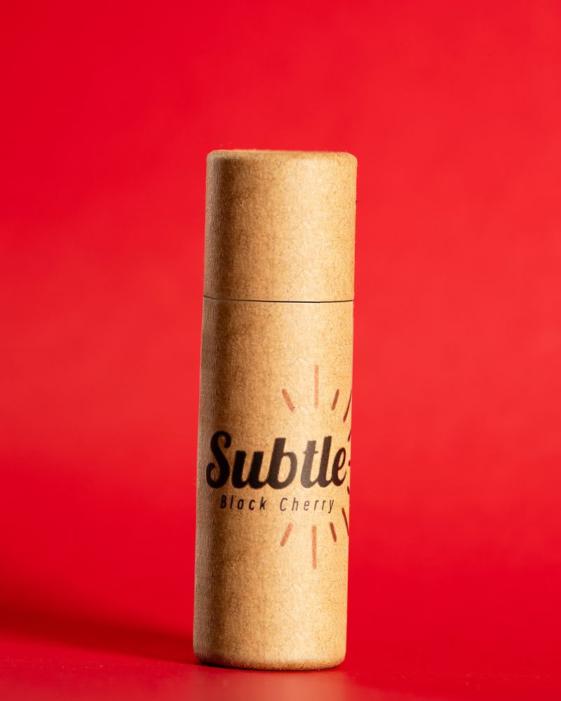 A kraft-paper Subtle lip balm tube standing in warm light against a red backdrop